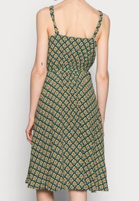 Grön blommig klänning med figurnära liv, justerbara axelband och vid kjol. Har ett mönster med upprepade orange och gula blommor.