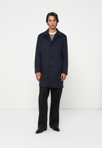 Uomo che indossa un cappotto blu marino con bottoni, un maglione a fantasia, pantaloni neri larghi e scarpe nere, in piedi contro uno sfondo bianco uniforme.