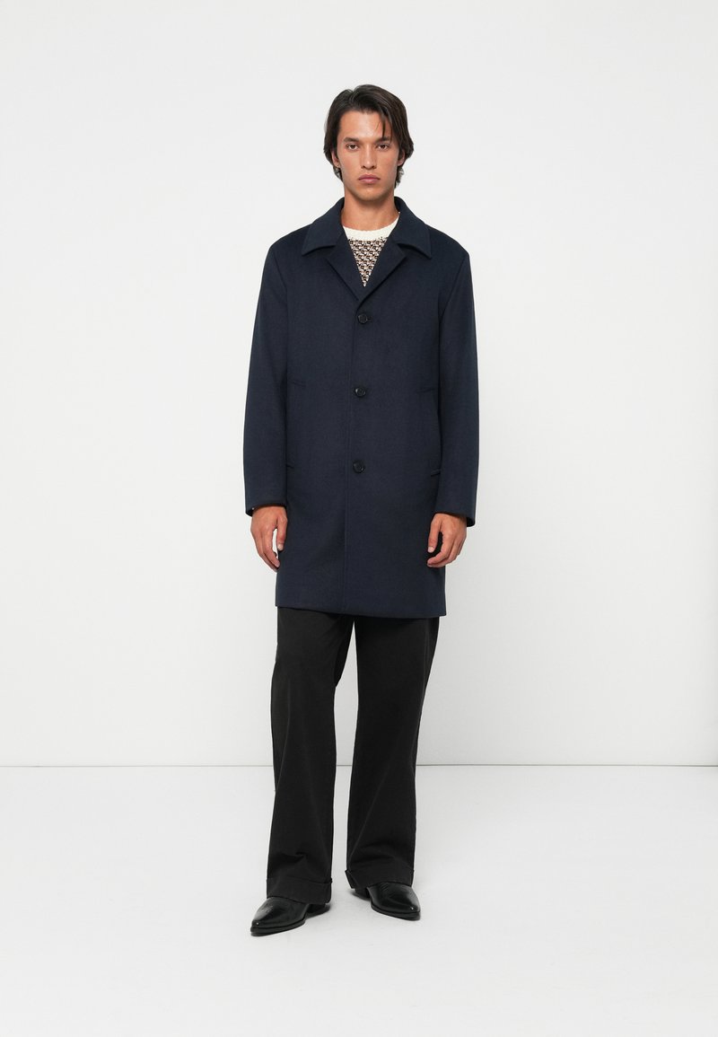 Uomo che indossa un cappotto blu marino con bottoni, un maglione a fantasia, pantaloni neri larghi e scarpe nere, in piedi contro uno sfondo bianco uniforme.