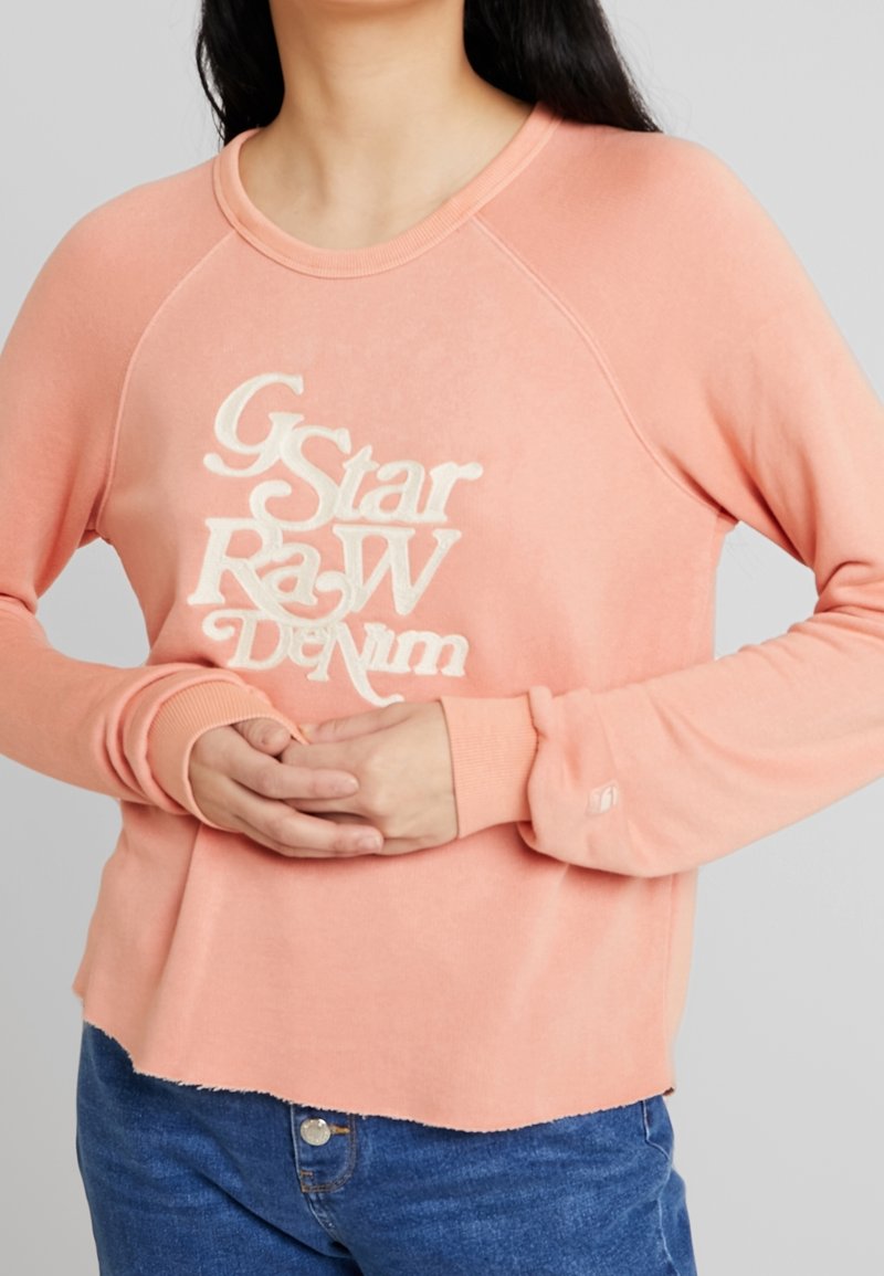 Sudadera rosa con diseño raglán, que presenta el texto bordado en blanco "G-Star Raw Denim"; puños y dobladillo acanalados, textura suave.