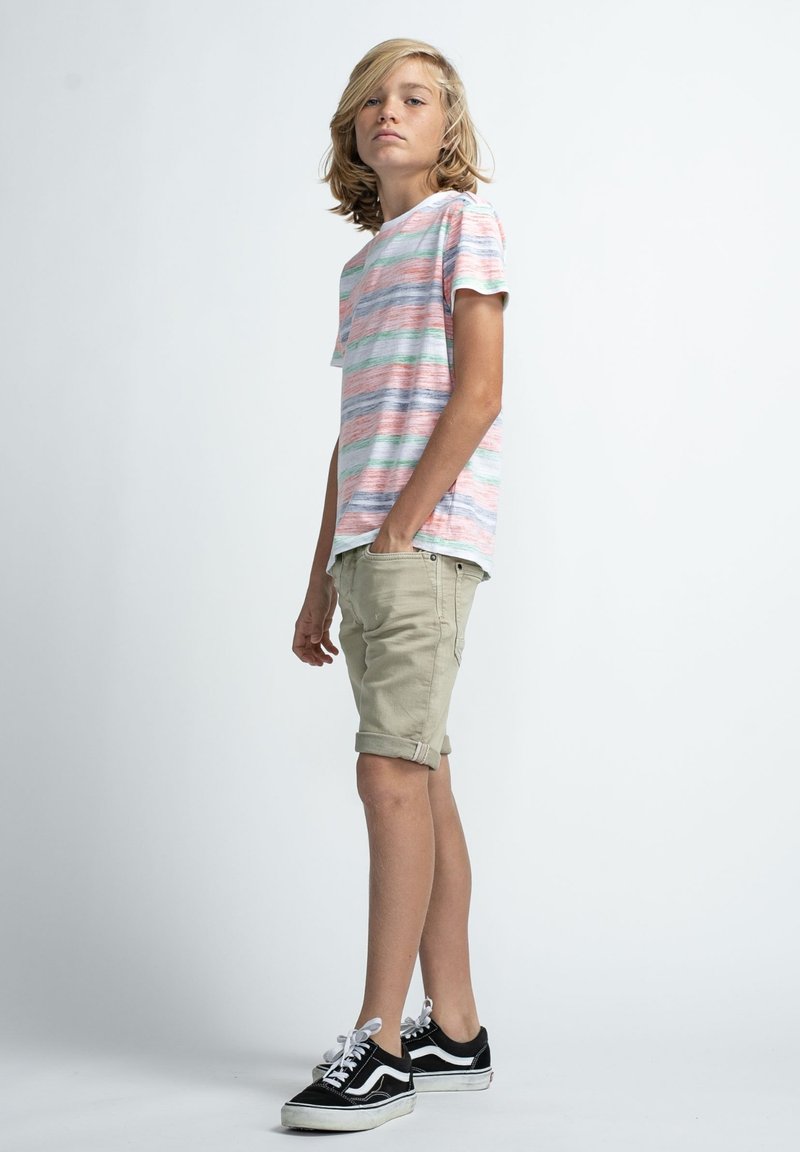 Gestreept t-shirt in roze, groen en blauw; beige opgerolde shorts; zwart-witte sneakers. Staande pose tegen een effen achtergrond.