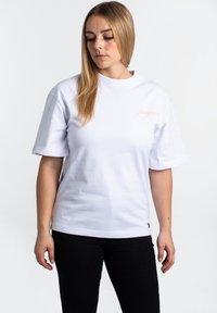 White cotton t-shirt with short sleeves, round neckline, featuring pink "frequ" text on the left chest area, paired with dark denim jeans.