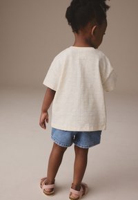 Lys gul kortærmet skjorte med blåt ternet mønster, kombineret med denimshorts. Barnet har på rosa sandaler og vender væk fra kameraet.