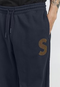 Marineblå joggebukser med elastisk midje, sidelomme og stort brunt "S"-logo på venstre lår. Myk stofftekstur.
