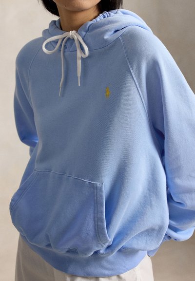 Personne portant un sweat à capuche bleu clair avec des cordons blancs et un logo brodé jaune sur le côté gauche de la poitrine, les mains dans la poche avant.