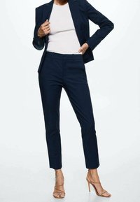 Costume pantalon bleu marine avec une veste cintrée et un pantalon fuselé. Tissu lisse, coupe sur mesure, assorti à des talons nus à brides.