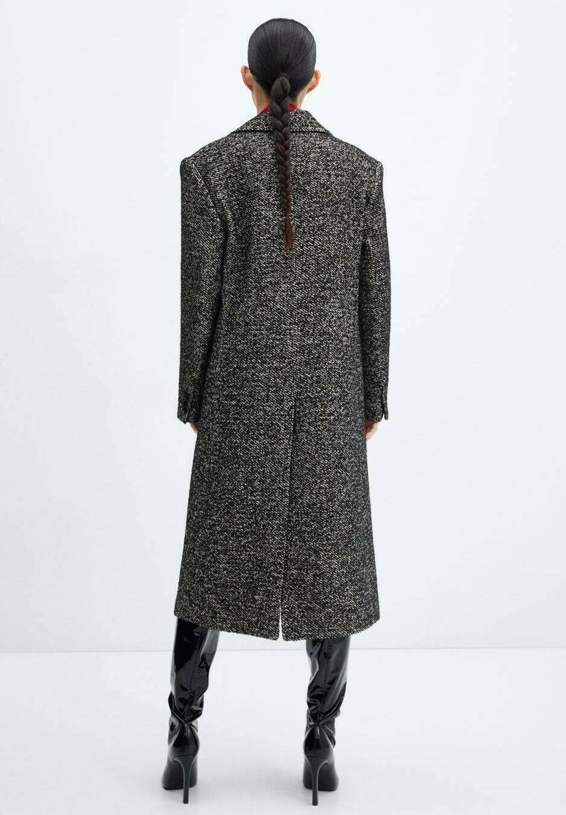 Un lungo cappotto strutturato in bianco e nero presenta spalle ampie, uno spacco sul retro e viene abbinato a stivali glossy neri alti fino alla coscia.