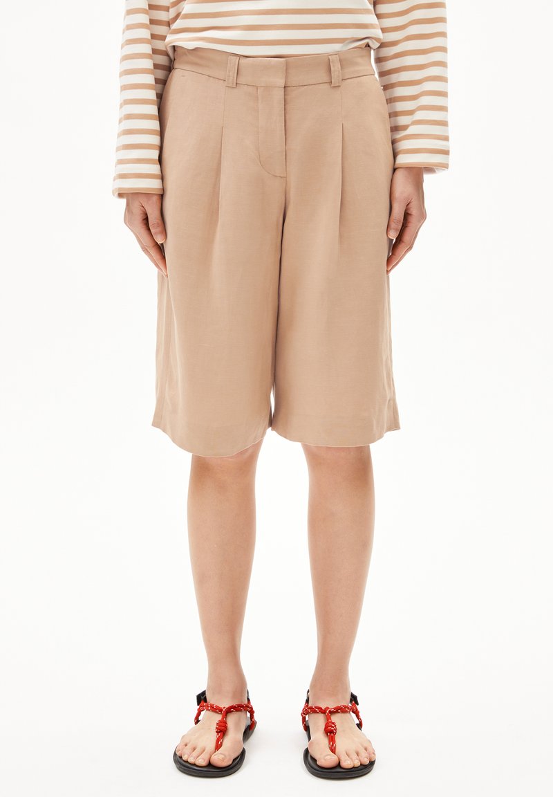 Beige Leinen-Shorts mit Seitenfalten, in lässiger Passform und mittellanger Oberschenkelhöhe, kombiniert mit schwarzen Sandalen mit roten Schnürelementen.