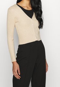 Beige ribbed cardigan with long sleeves and a V-neck, featuring small buttons down the front and a slightly cropped design.