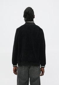 Homem com dreadlocks vestindo um casaco preto de veludo cotelê e jeans pretos desbotados, de costas, sobre um fundo branco liso.