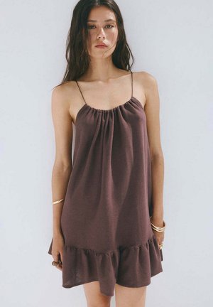 Mujer con cabello largo y oscuro que lleva un vestido marrón suelto sin mangas con tirantes finos y dobladillo fruncido, de pie contra un fondo claro.