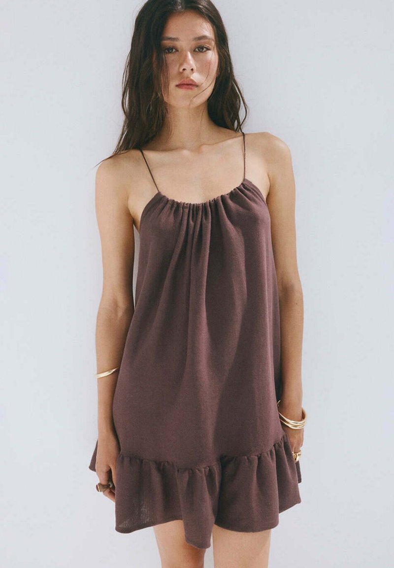Femme aux longs cheveux foncés portant une robe marron ample, sans manches, avec des fines bretelles et un ourlet à volants, debout devant un fond clair.