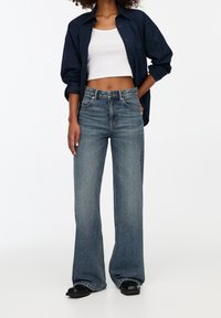 Vaaleat siniset leveälahkeiset farkut, joissa on haalistunut viimeistely, yhdistettynä valkoiseen crop-toppiin ja tummansiniseen napitettuun paitaan. Mustat kengät viimeistelevät asun.