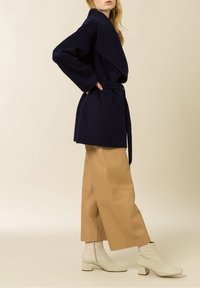 Abrigo envolvente de lana azul marino con mangas anchas y cinturón, combinado con pantalones de pierna ancha de cuero marrón claro y botas de tobillo beige con relieve de cocodrilo.