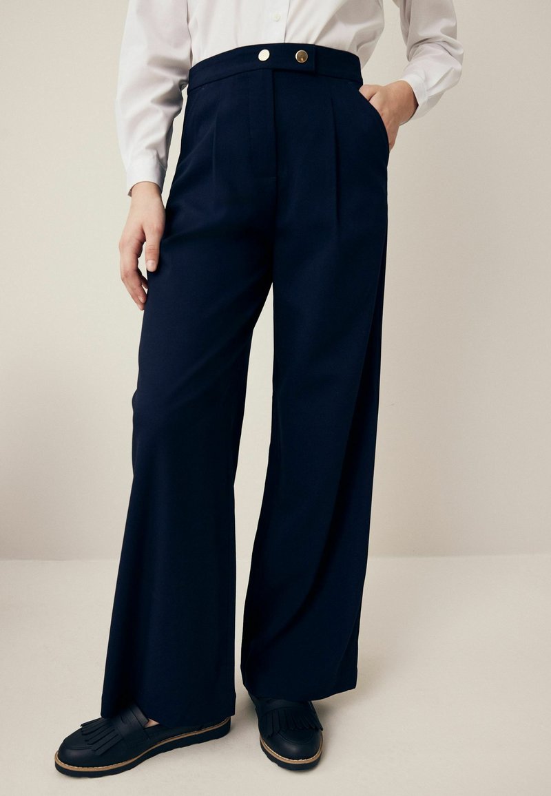 Tamsiai mėlyni platėjančių kelnių modelis su aukšta juosmeniu, dviem priekinėmis kišenėmis ir auksiniu sagos dekoru. Audinys atrodo lygus ir struktūruotas.