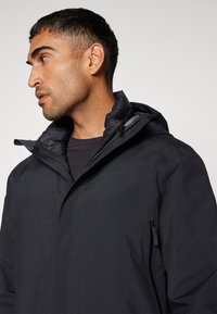 Chaqueta negra impermeable con capucha, cierre de cremallera y un interior texturizado, que cuenta con un bolsillo lateral y un diseño elegante y moderno.