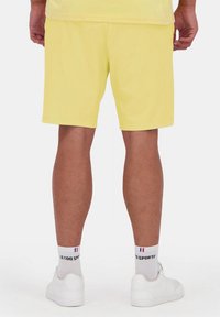 Pantalones cortos deportivos amarillos hechos de una tela suave, de longitud hasta la rodilla con una textura lisa, combinados con calcetas deportivas blancas y zapatillas blancas.