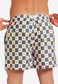 Costume da bagno con una base blu caratterizzato da un motivo geometrico di quadrati bianchi, cerchi verdi e diamanti arancioni. Vita elastica, tasche laterali.