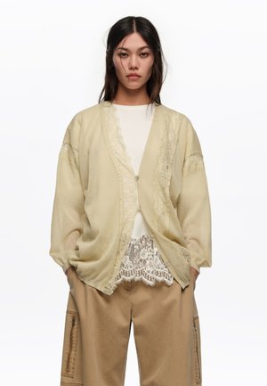 Mujer con un cárdigan beige con ribete de encaje sobre una blusa blanca de encaje y pantalones cargo sueltos color marrón claro, de pie con las manos en los bolsillos contra un fondo blanco.