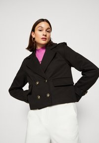 Czarna dwurzędowa marynarka z złotymi guzikami, kieszeniami naszytymi i naramiennikami. Tkaninowy tekstur i krótki fason.