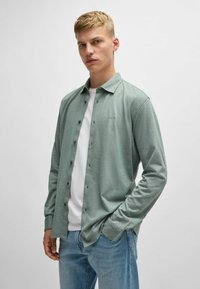 Lichtgroen overhemd met lange mouwen en knoopjes, met een kraag, voorzien van een subtiel logo, gedragen over een wit t-shirt, gecombineerd met een blauwe jeans.