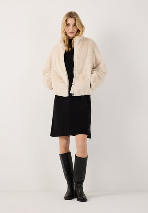 Frau trägt eine beige Kunstpelzjacke über einem schwarzen Kleid mit kniehohen schwarzen Lederstiefeln und steht vor einem schlichten Hintergrund.