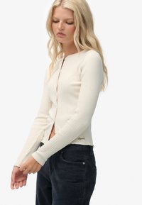 Prążkowany kardigan z długim rękawem w kolorze off-white z okrągłym dekoltem, zapięciem na guziki z przodu i krótkim dołem. Zawiera złote logo po boku.