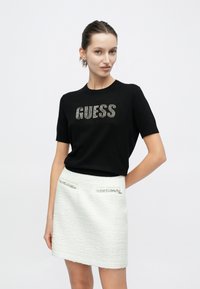 Schwarzes T-Shirt mit kurzen Ärmeln und der Aufschrift "GUESS" in verzierten Buchstaben, kombiniert mit einem weißen, strukturierten Minirock mit zwei dekorativen Taschen.