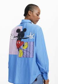 Blauw gestreept shirt met lange mouwen. De achterkant heeft een grote afbeelding van Mickey Mouse, in zwart en rood, tegen kleurrijke patchworkpanelen.