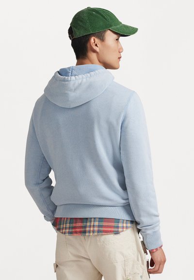 Ανοιχτό μπλε φούτερ με κουκούλα, ριμπ μανσέτες και στρίφωμα, φορεμένο πάνω από ένα κόκκινο και πράσινο καρό πουκάμισο. Φοριέται με ένα πράσινο καπέλο από κοτλέ.