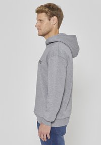 Hall hallitus tehtud pehmest kangast, millel on kängurutasku ja reguleeritava paelaga kapuuts. Modell kannab siniseid teksapükse, näidatud küljelt.