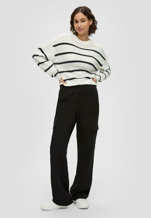 Witte gebreide trui met zwarte horizontale strepen, gecombineerd met zwarte cargo broek met dubbele zijzakken, afgewerkt met witte sneakers.