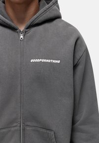 Hoodie zippé gris en tissu doux. Il possède une poche kangourou et le texte blanc "GOODFORNOTHING" sur la partie gauche de la poitrine.