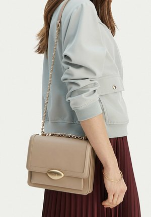 Kobieta ubrana w jasnoniebieską kurtkę i bordo spódnicę trzyma na ramieniu beżową torbę na ramię ze złotym łańcuszkiem i zapięciem.