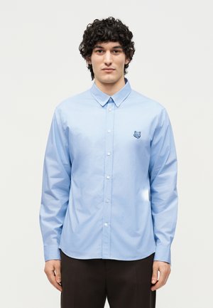 Man with curly black hair wearing a light blue button-up shirt with a small embroidered animal logo on the left chest, standing against white background.