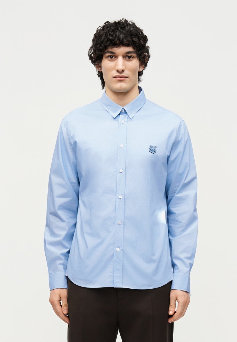 Άνδρας με σγουρά μαύρα μαλλιά που φοράει ένα ανοιχτό γαλάζιο πουκάμισο με κουμπιά και μικρό κεντητό λογότυπο ζώου στο αριστερό στήθος, στέκεται μπροστά σε λευκό φόντο.