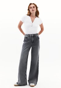 Vaqueros anchos negros de cintura alta, con una tela de mezclilla texturizada y bolsillos delanteros. Combinados con una blusa ajustada de manga corta blanca.
