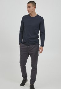 Marineblauw shirt met lange mouwen, gecombineerd met donkergrijze cargo-broeken met meerdere zakken. Zwarte sneakers met witte zolen maken de outfit compleet.