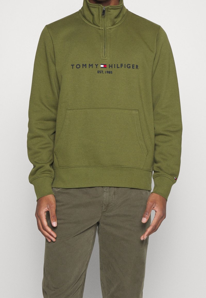 Homme portant un sweat-shirt Tommy Hilfiger à demi-fermeture éclair couleur vert olive avec une poche avant et un pantalon assorti vert olive.