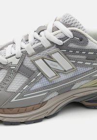 Zbliżenie na szare i białe siatkowe sneakersy z białymi sznurówkami i logo "N" po boku, podkreślające szczegółowe przeszycia i wzór podeszwy.