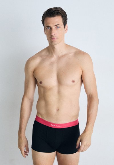 Fekete boxeralsó rózsaszín derékszallaggal. A derékszallagon a "Paul Smith" logó található. A modell mezítelen felső testtel áll egy világos háttér előtt.