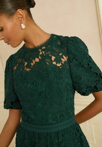 Smaržīga zaļā mežģīņu kleita ar pūkainām piedurknēm, dekorēta ar ziedu rakstiem, ar viļņotu apkakli un pieguļošu vidukli ar mežģīnēm.