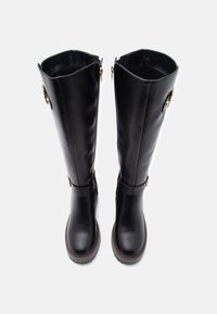 Bottes noires en cuir montantes avec une texture lisse, zips latéraux et une semelle épaisse marron, dotées d'une sangle et de détails en métal doré.