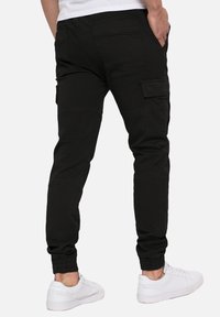 Pantalon cargo noir en tissu robuste, avec plusieurs poches, coupe ajustée, poignets côtelés et texture lisse. Stylé avec des baskets blanches.