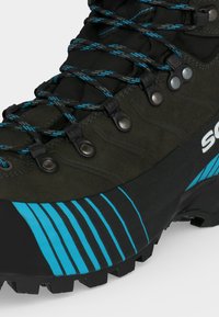 Bota de senderismo verde oscura con suela a rayas azul y negra, cordones negros y azules, ojales de metal y suela resistente para uso al aire libre.