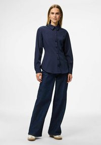 Woman wearing a navy blue pinstripe button-up shirt, wide-leg dark blue jeans, and beige sneakers, standing against a plain white background.