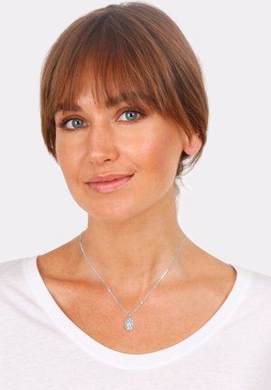 Silberne Halskette mit einem kleinen Anhänger, getragen über einem weißen Hemd. Der Anhänger ist mit feinen Gravuren versehen; die Kette ist schlank und glatt.