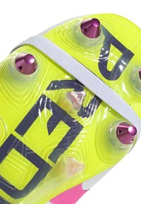 Crampon jaune vif avec un dessus transparent, des accents roses, des crampons métalliques violets et une sangle blanche. Présente de grandes lettres noires sur le côté.