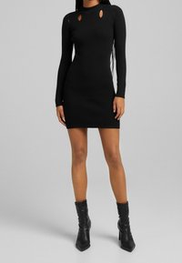Svart ribbad stickad klänning med långa ärmar, med utskärningar vid axlarna och en åtsittande siluett, kombinerad med svarta klackstövlar.