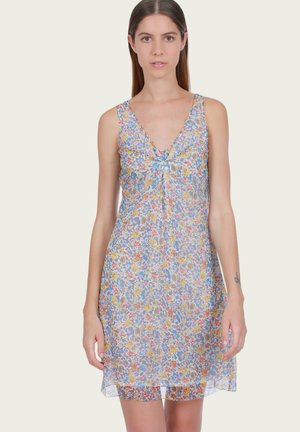 Femme portant une robe à fleurs sans manches avec col en V et détail noué, debout devant un fond clair uni.